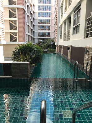 A swimming pool with clear blue water, surrounded by a modern building with multiple floors and balconies. - The Link Onnut by May (Bangkok)