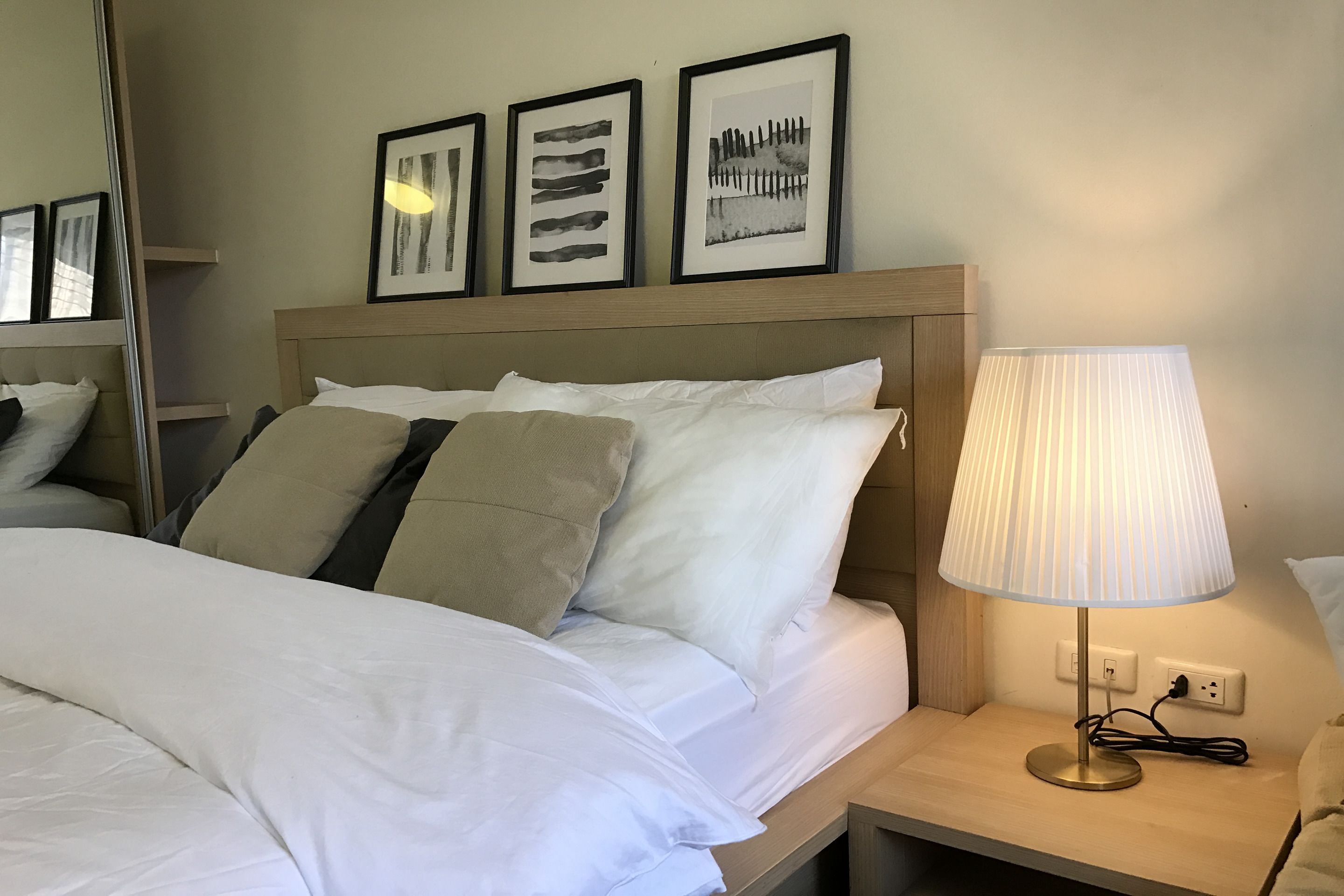 A neatly made bed with white and beige bedding, a bedside lamp, and three framed artworks on the wall.