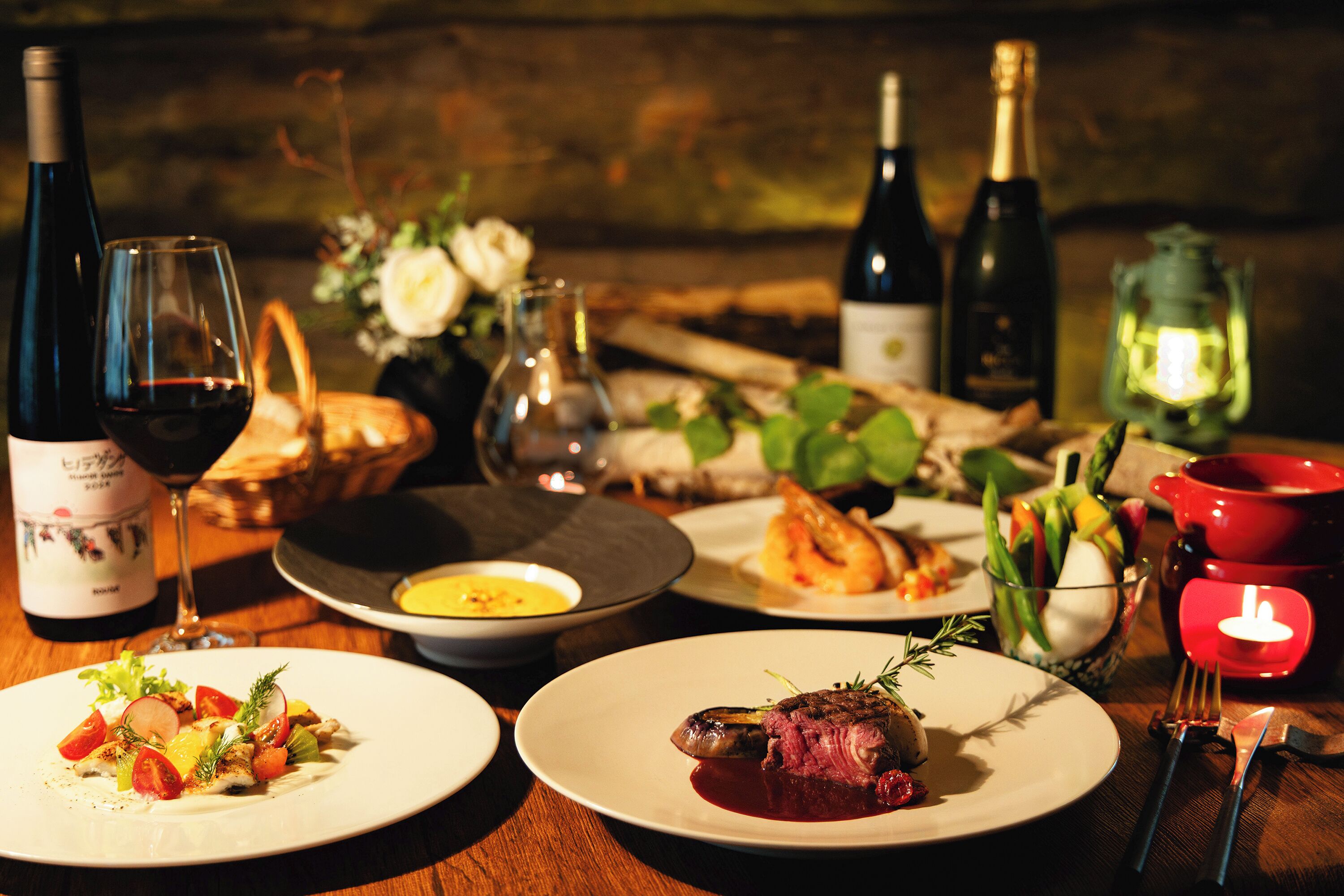 A dining table with plates of food, wine bottles, glasses, and a lit lantern.