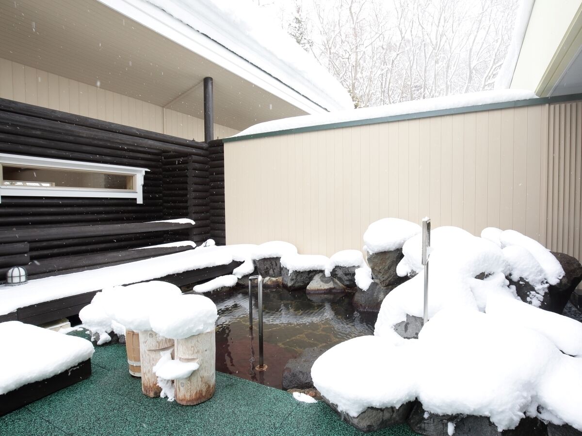 A snow-covered outdoor area with a hot spring, wooden benches, and a stone structure.