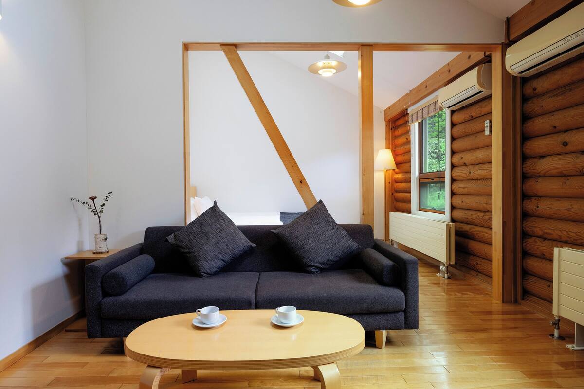 Una habitación con un sofá oscuro, una mesa redonda de madera con dos tazas, suelo de madera y una pared de madera.