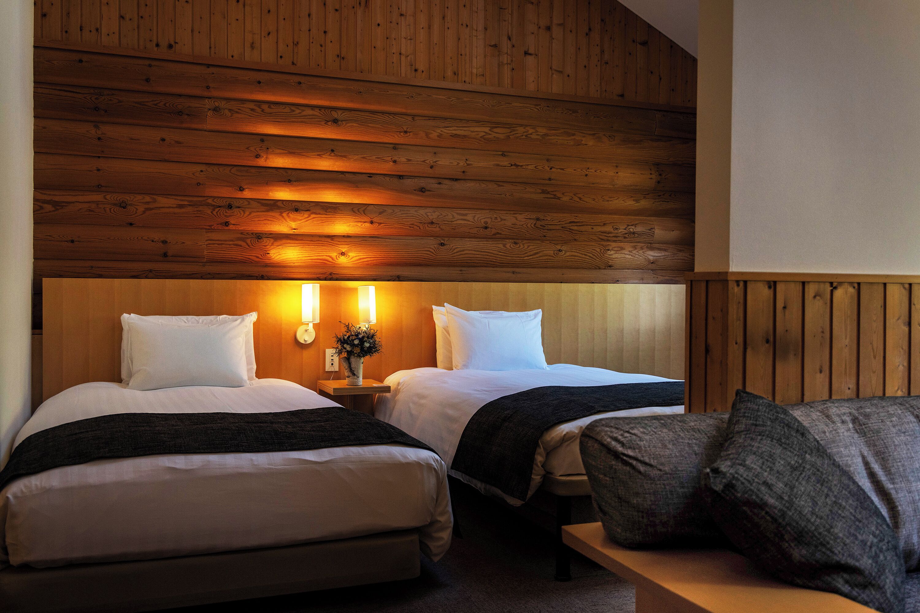 Ein Hotelzimmer mit zwei Betten, Holzzapfen an der Decke, einem Nachttisch mit Blumenvase und einer Bank mit Kissen.
