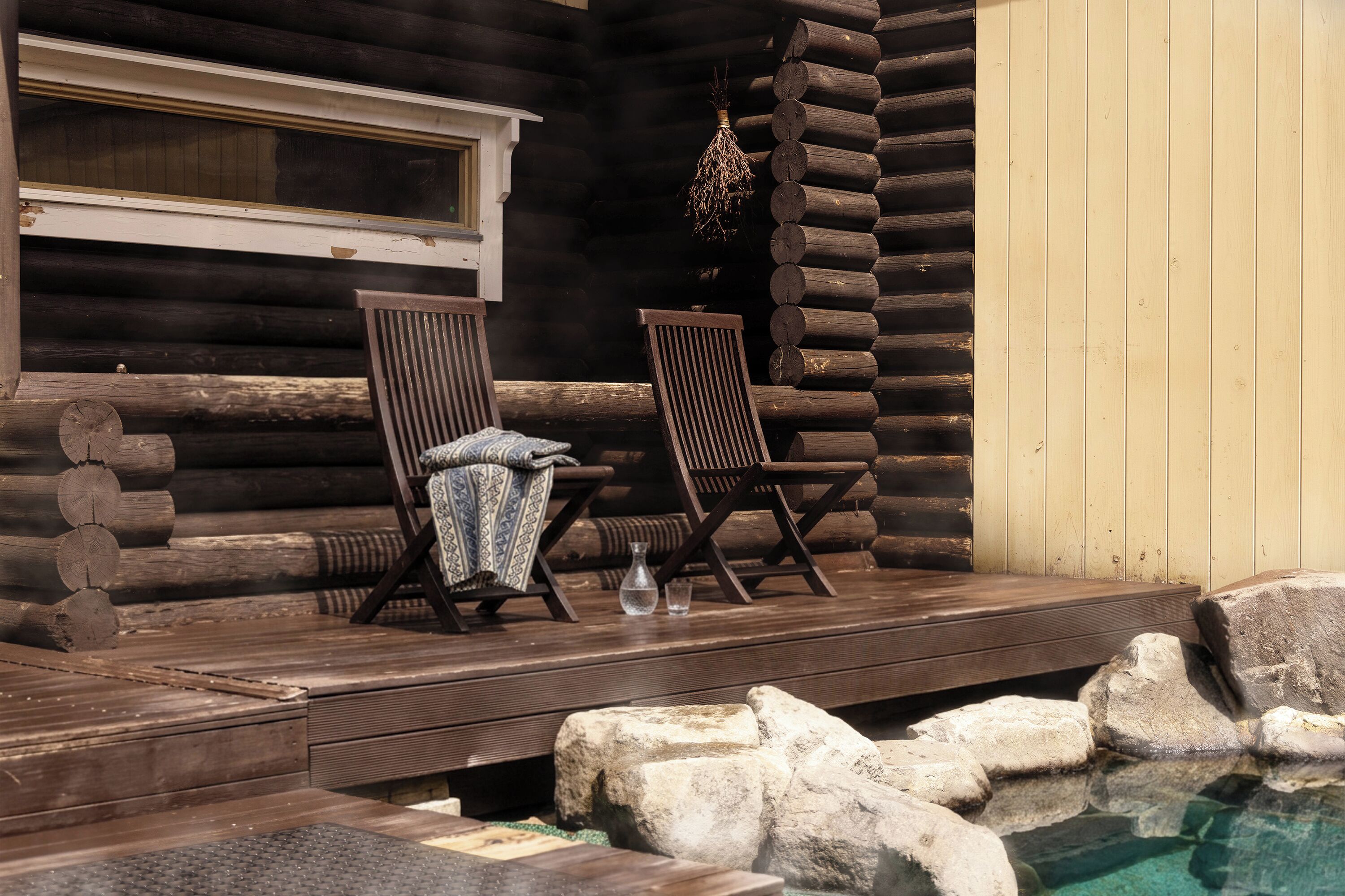 A log cabin with a wooden deck, two wooden chairs, a small table, and a clear pool with rocks.