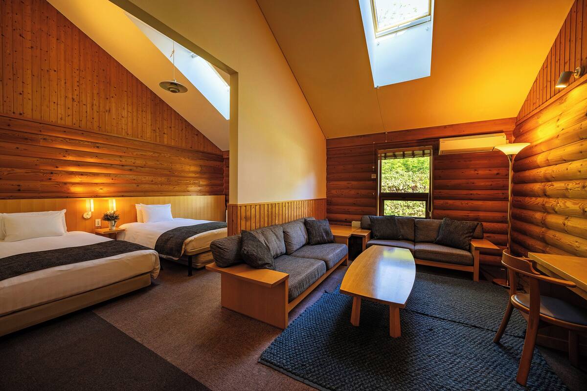 Interior de una cabaña de troncos con un dormitorio en la superior, un sofá, una mesa de comedor y una lámpara.