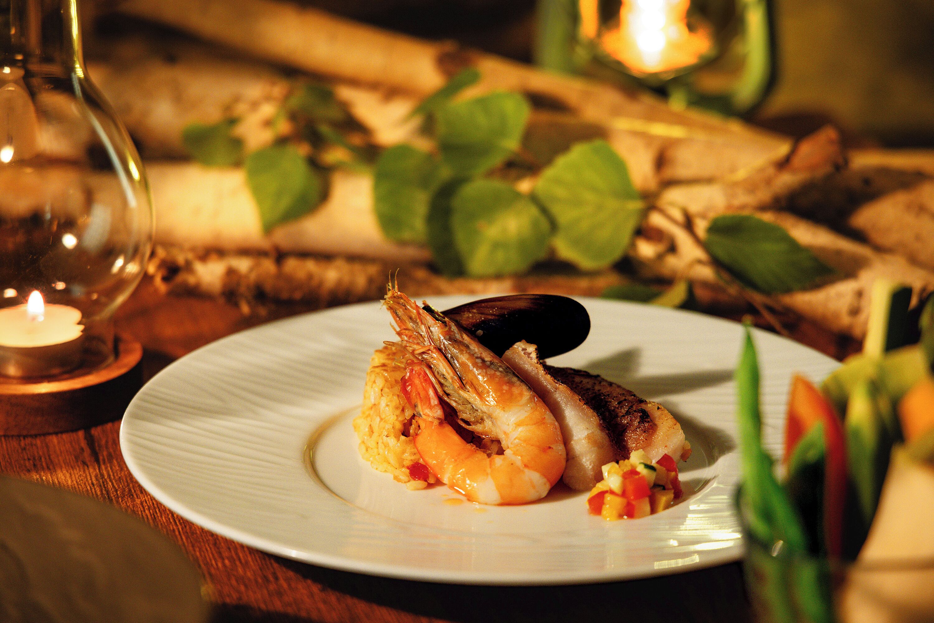 A plate with seafood and vegetables on a wooden table, surrounded by greenery and a lit candle.
