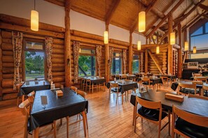 A log cabin-style restaurant with wooden tables and chairs, large windows, and hanging pendant lights.