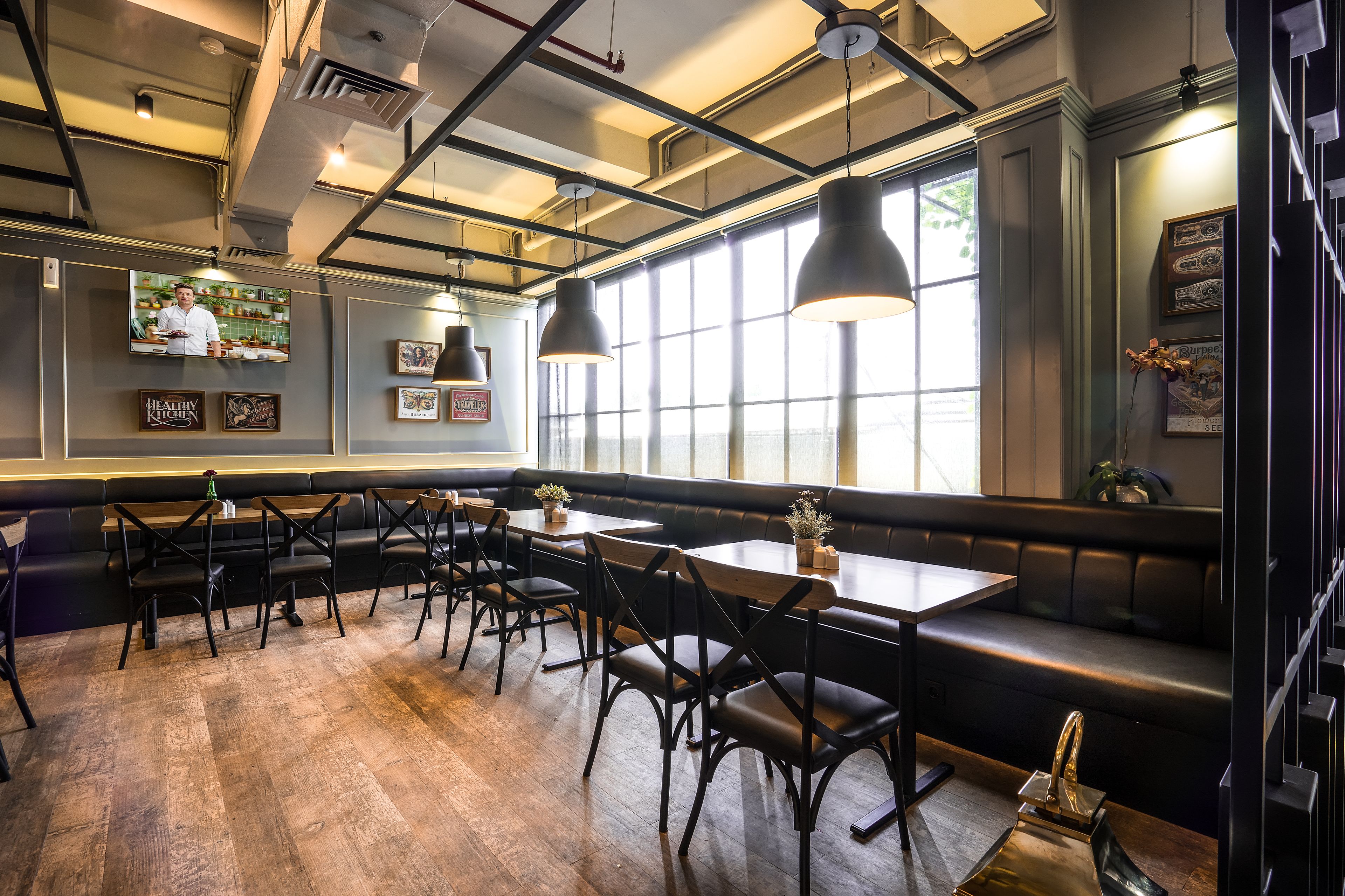 A restaurant interior with wooden flooring, black leather seating, pendant lighting, and a television displaying a chef.