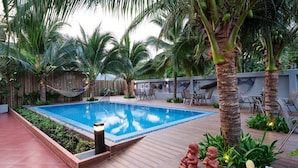Piscina rectangular con muelle de madera, palmeras y mobiliario exterior.