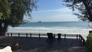 A wooden deck with chairs and tables overlooking a beach with waves and a distant island. - Away Las Tortugas Beachfront (Hua Hin)