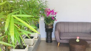 A living room with a grey sofa, a wicker side table, and a potted plant with pink flowers. - Kavil Guesthouse (Chiang Mai)