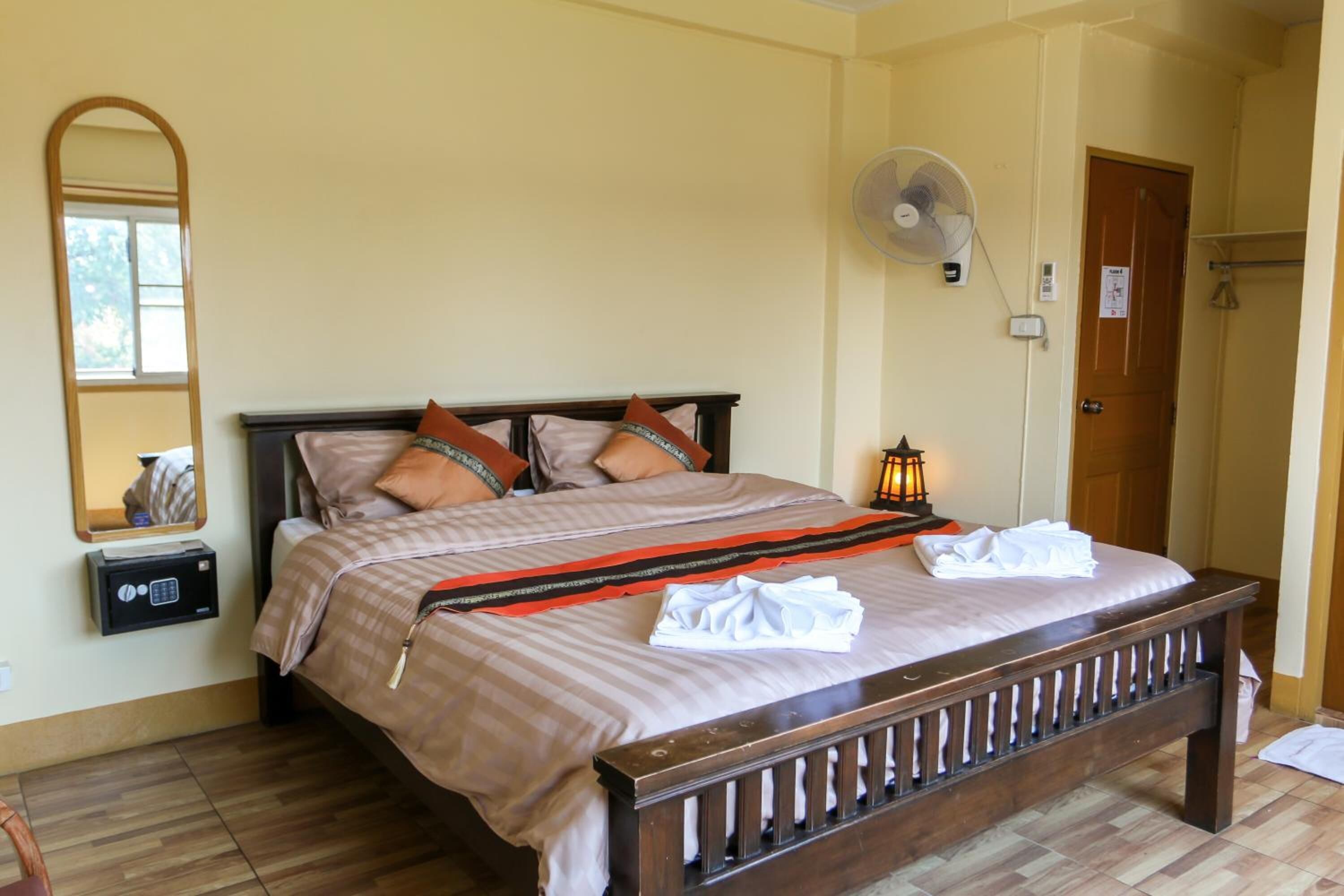 A hotel room with a bed, a bedside table with a safe, a wall-mounted fan, and a door with a towel rack.