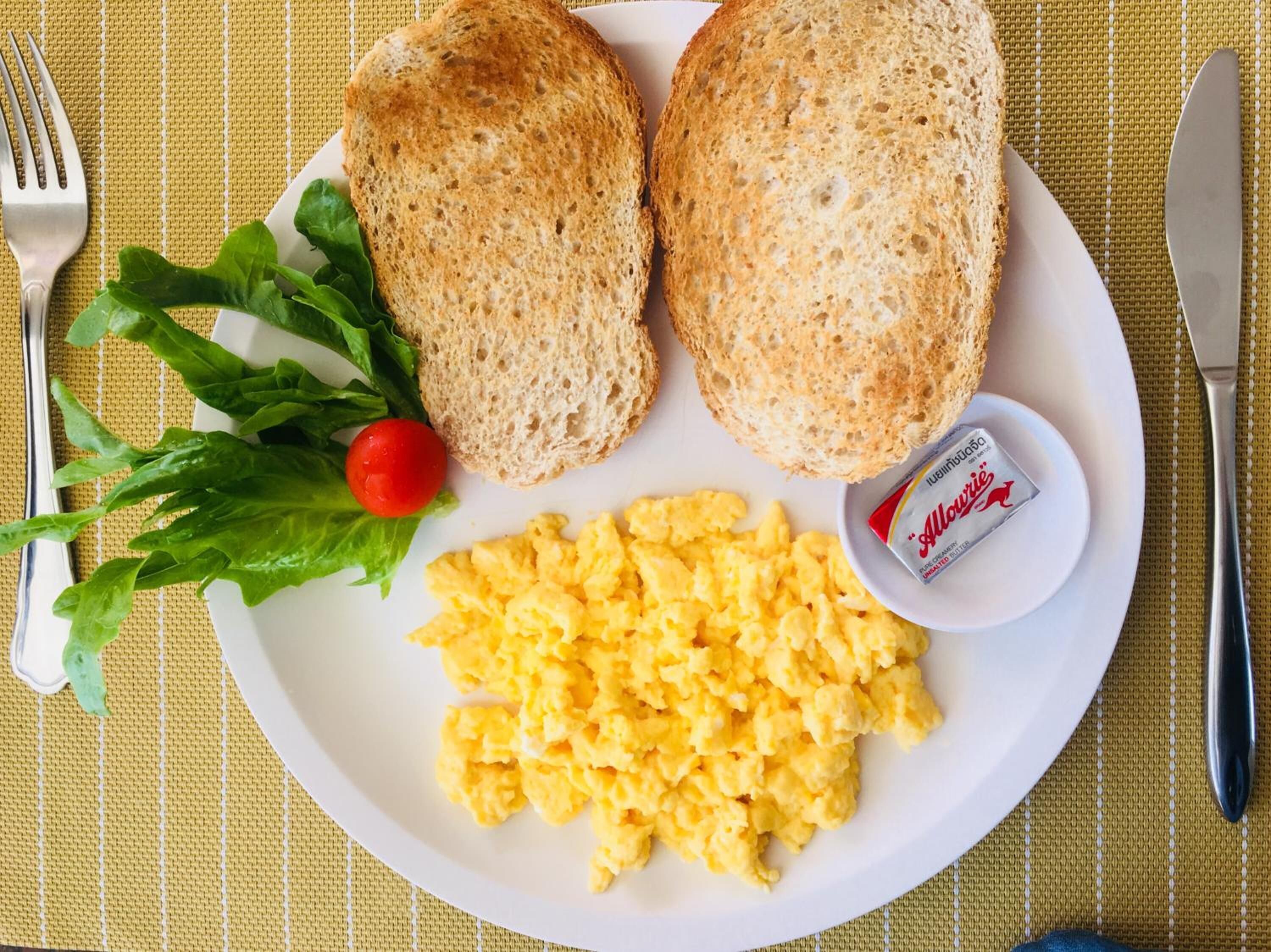 A plate with scrambled eggs, two slices of bread, a small container of butter, a cherry tomato, and some greens.