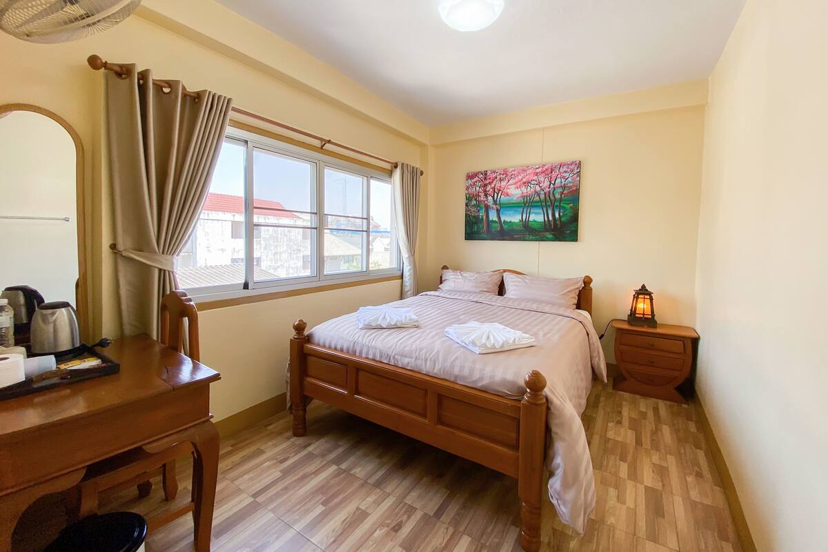 A bedroom with a wooden bed, a desk with a chair, a window with curtains, a painting on the wall, and a nightstand with a lamp.