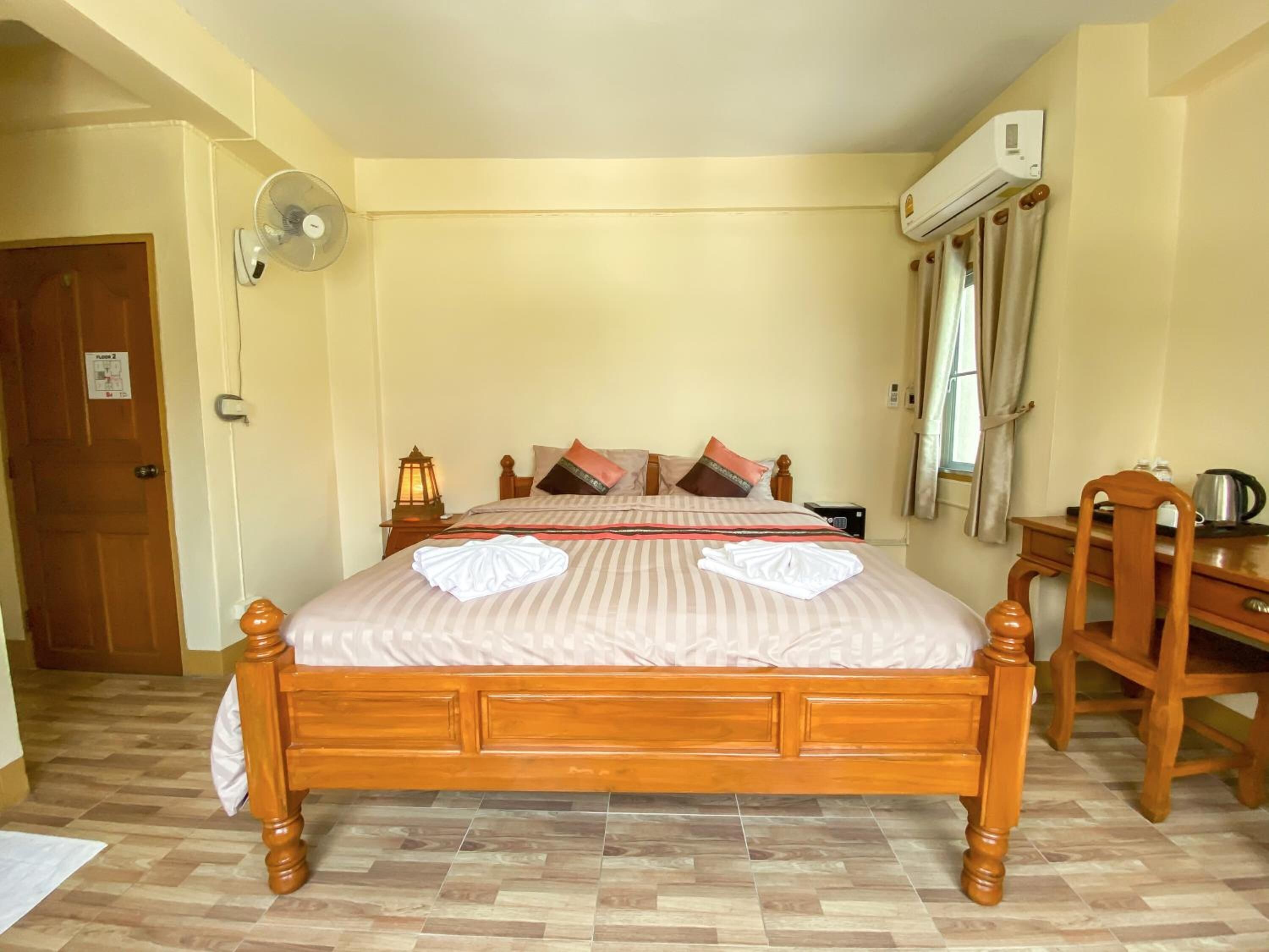 A hotel room with a wooden bed, a desk, a chair, a fan, and an air conditioning unit.