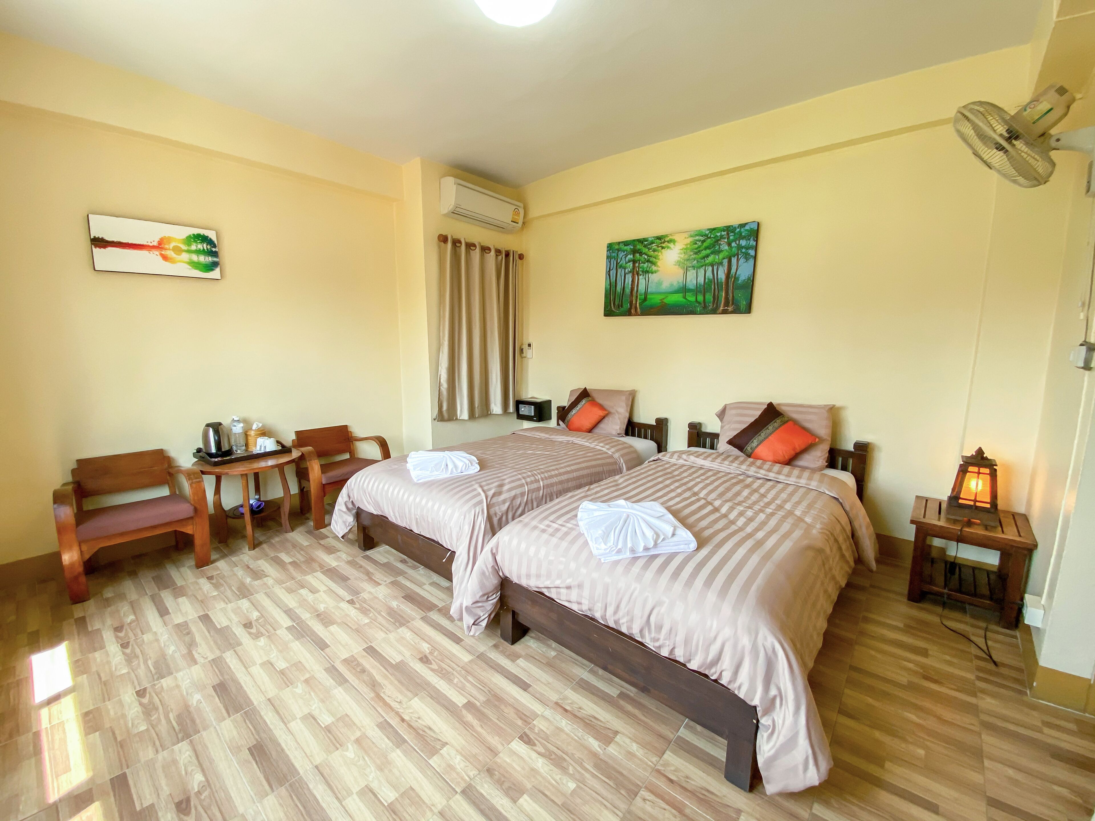 A hotel room with two beds, a wooden desk, a chair, a table with a kettle, a fan, and a painting on the wall.