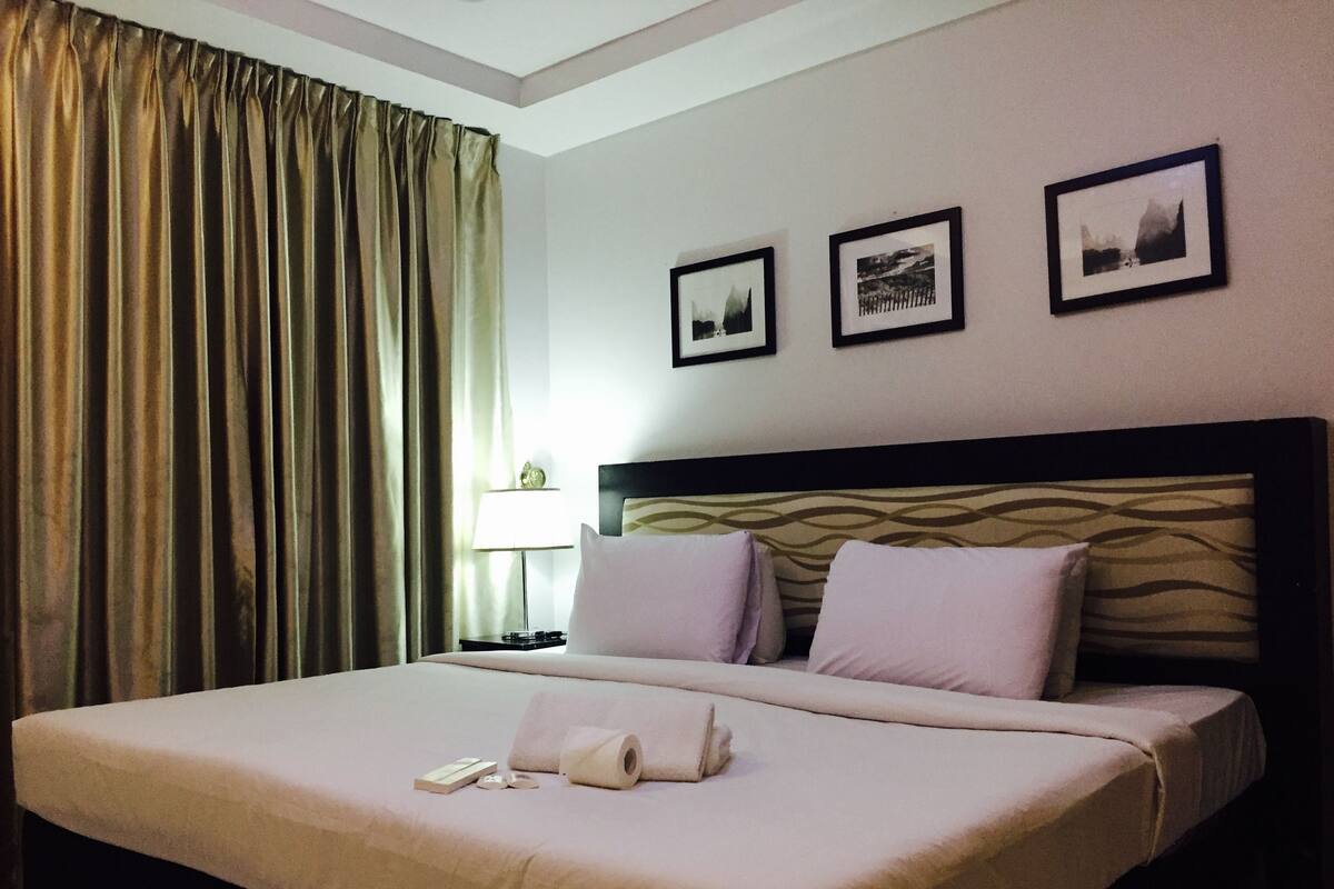 Hotellihuone, jossa on suuri sänky, kaksi tyynyä, yöpöytä lampun kanssa ja kolme kehystettyä kuvaa seinällä.