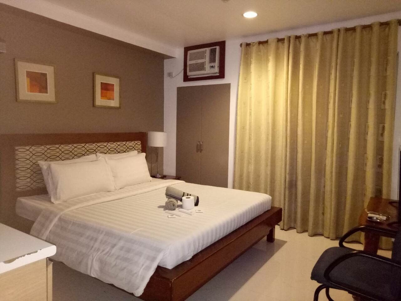 Quarto de hotel com uma cama grande, uma escrivaninha, uma cadeira, um guarda-roupa e uma janela com cortinas.