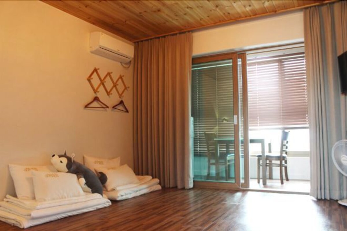 A room with a wooden ceiling, a bed with white linens, a stuffed toy, a wall-mounted air conditioner, and a sliding glass door leading to an outdoor area with chairs.