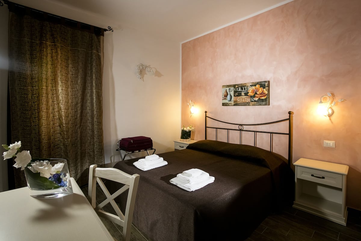 Una cama bien hecha con una colcha marrón, dos toallas blancas, un jarrón con flores, una maleta roja y una pared con un letrero decorativo.