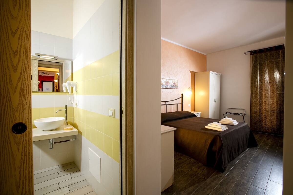 Un baño con lavamanos, espejo y barra para toallas, y un dormitorio con cama, mesita de noche y armario.