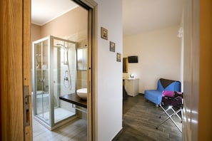 A modern bathroom with a glass shower enclosure, a sink, and a toilet. There is a small kitchenette area with a microwave and a cabinet. A blue chair and a pink suitcase are visible in the room. - Triskell Camere & Relax (San Vito Lo Capo)