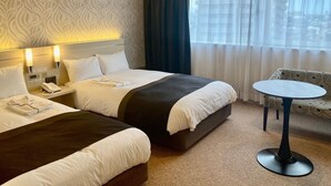 Une chambre d’hôtel avec deux lits, une table de chevet avec un téléphone, une petite table ronde et une vue sur la ville à travers de fines rideaux.