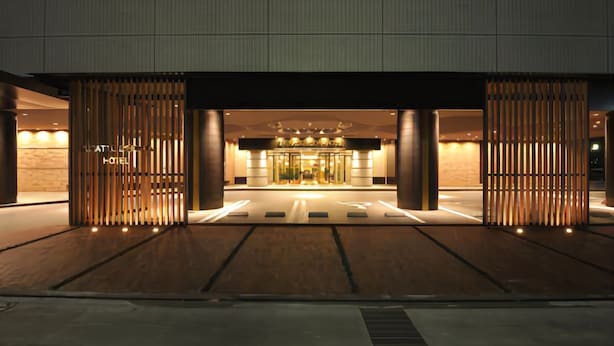 Une entrée d’hôtel moderne avec une façade en verre, des lambris en bois verticaux et un espace d’accueil bien éclairé.