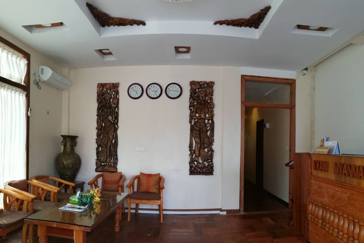 A spacious living room with a wooden dining table, two wooden chairs, a large vase, and a decorative wooden carving on the wall.
