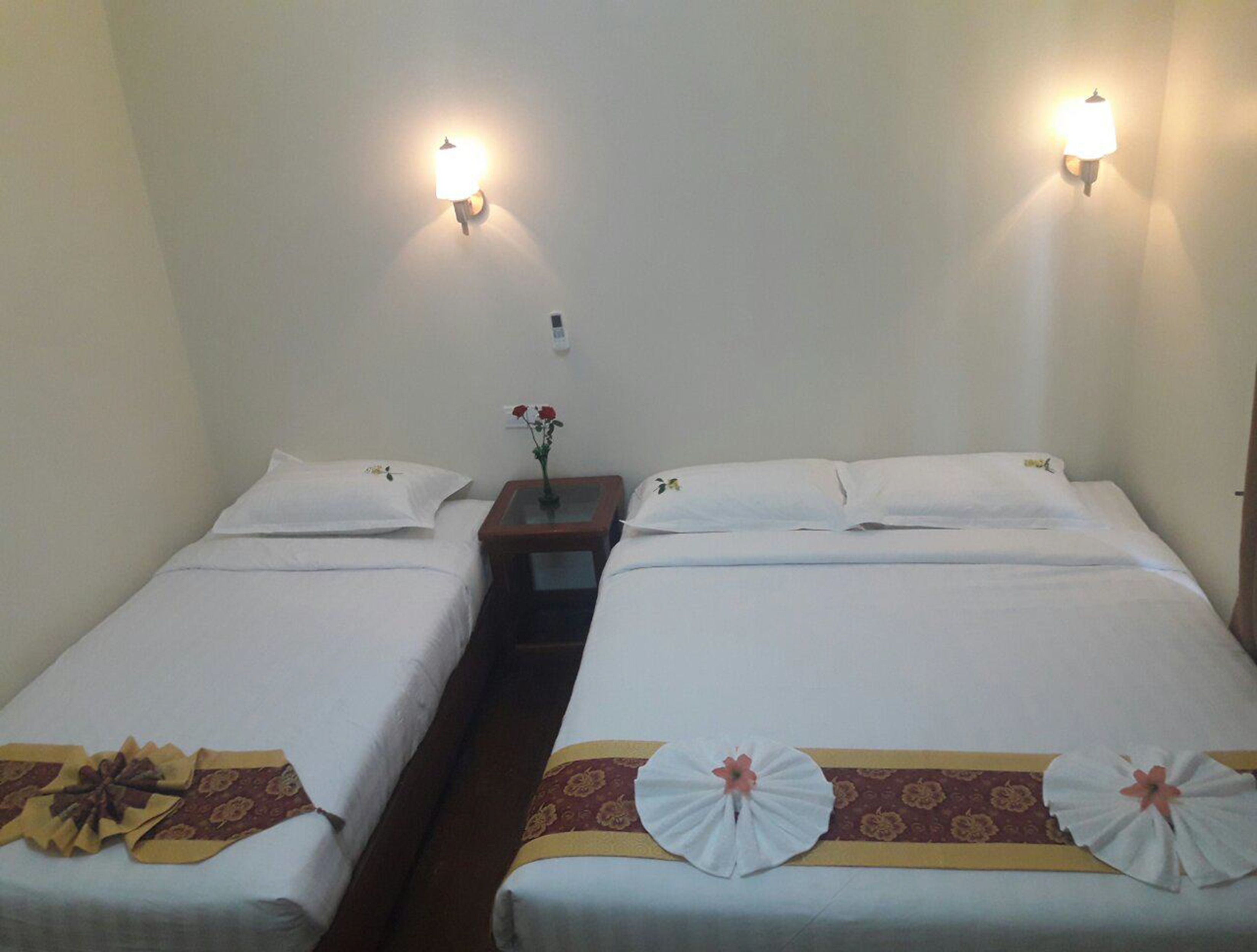 A hotel room with two single beds, each with a floral bedspread and a decorative pillow. There are two wall-mounted lamps and a small table with a vase of flowers between the beds.