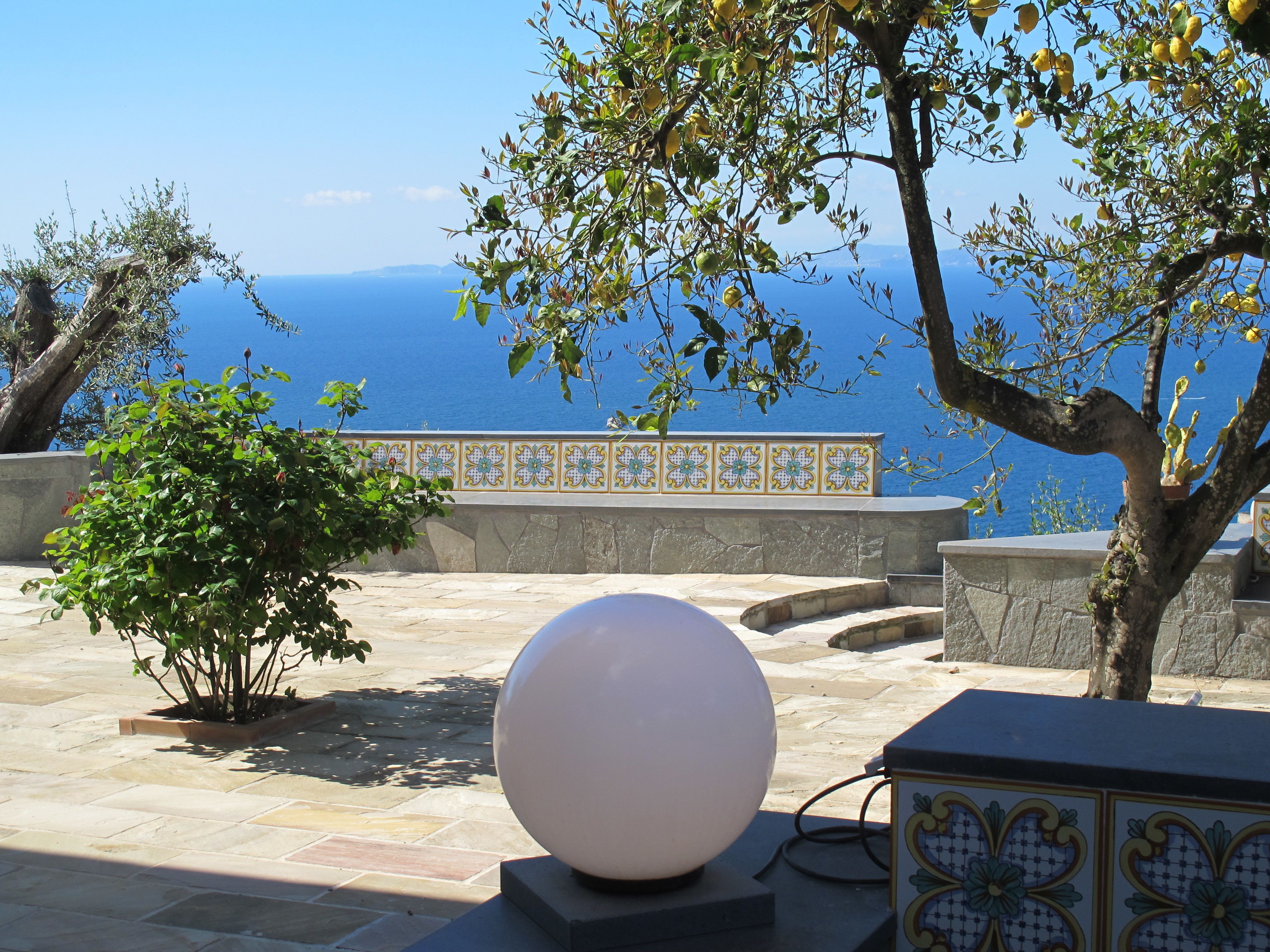 A stone pathway leading to a terrace with a view of the sea, a white spherical object, and a tiled wall with floral patterns.