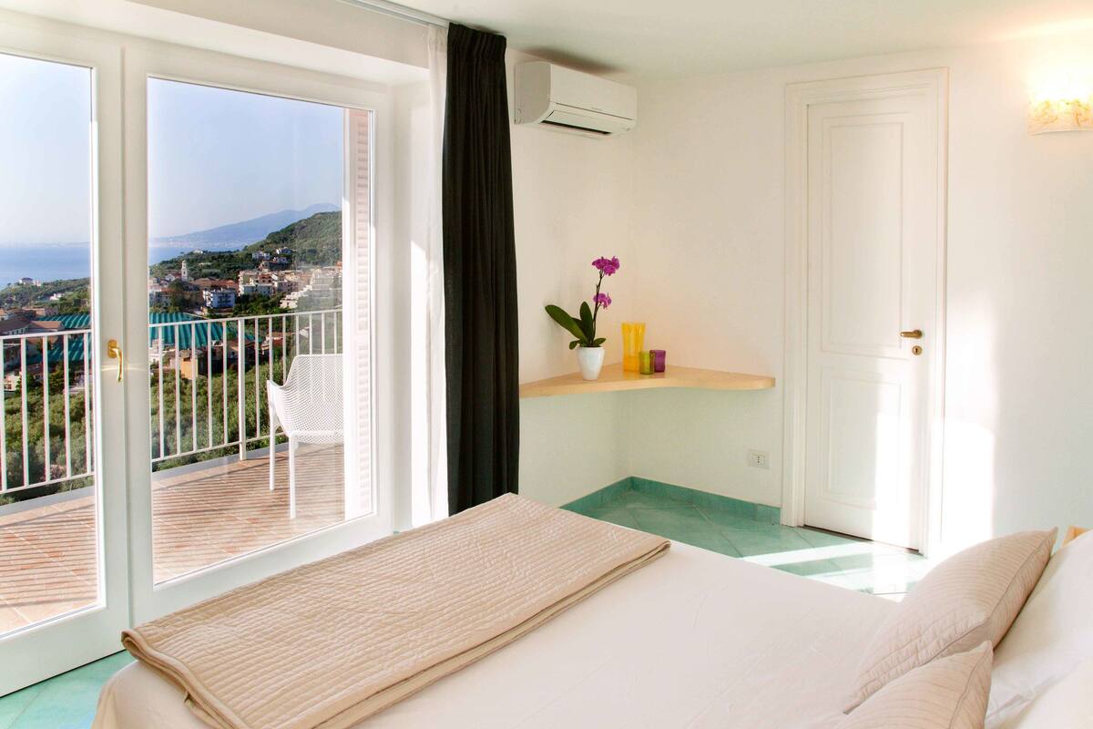 Une chambre d’hôtel avec un lit, une petite table sur laquelle est posée une plante en pot, un balcon avec une chaise, et une vue sur la mer et les bâtiments.