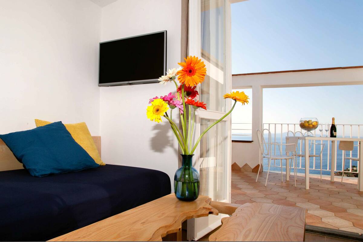 Une chambre avec un lit, un téléviseur fixé au mur, un vase avec des fleurs et un balcon avec une table et des chaises.