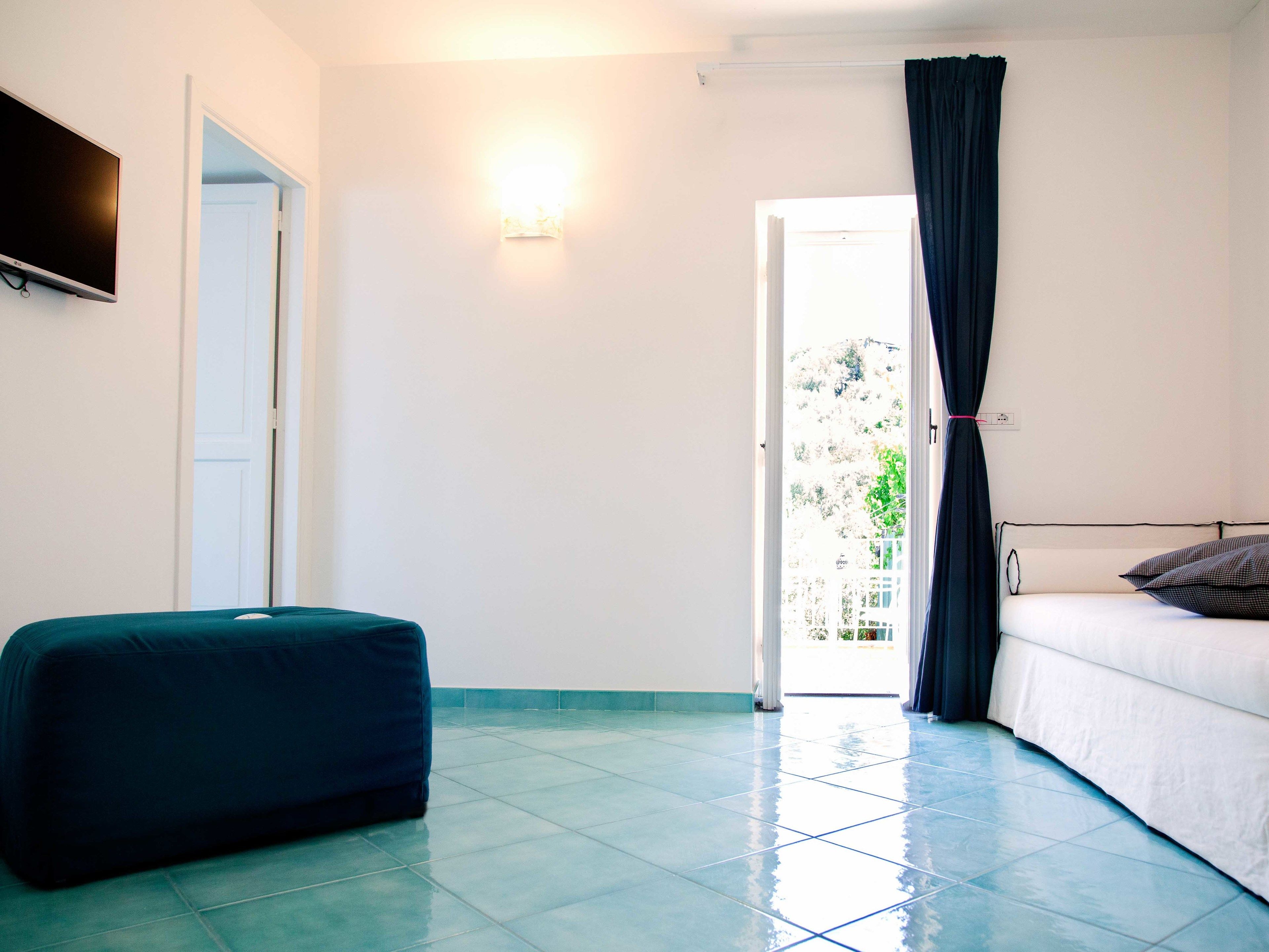 A room with a bed, a TV mounted on the wall, a window with curtains, and a tiled floor.