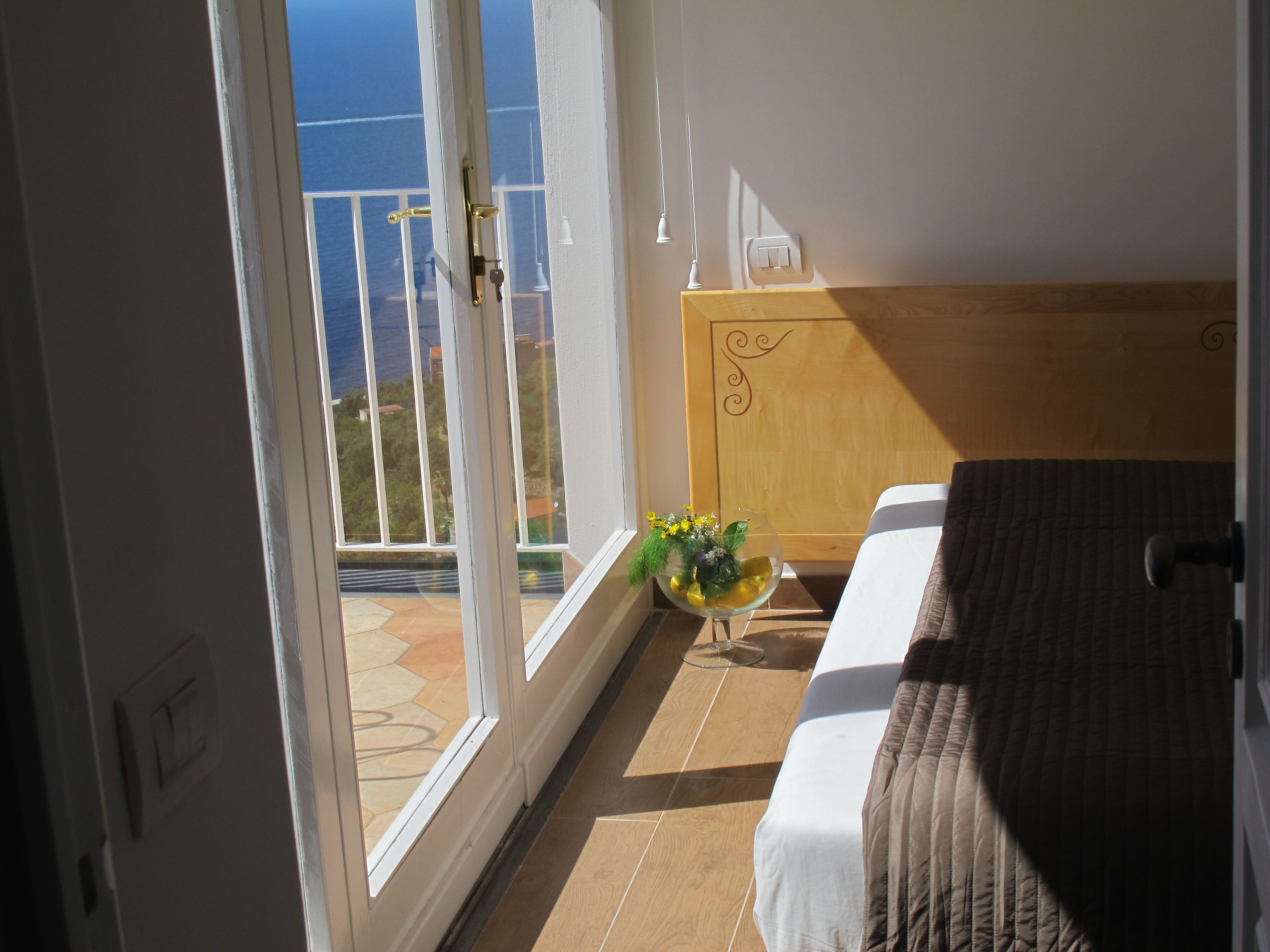 A bedroom with a bed, a wooden headboard, a small table with a vase of flowers, and a view of the sea through a sliding glass door.