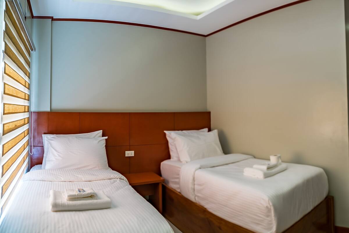 Een hotelkamer met twee bedden, houten hoofdeinden, wit beddengoed, een klein nachtkastje met een lamp, en een houten deur.