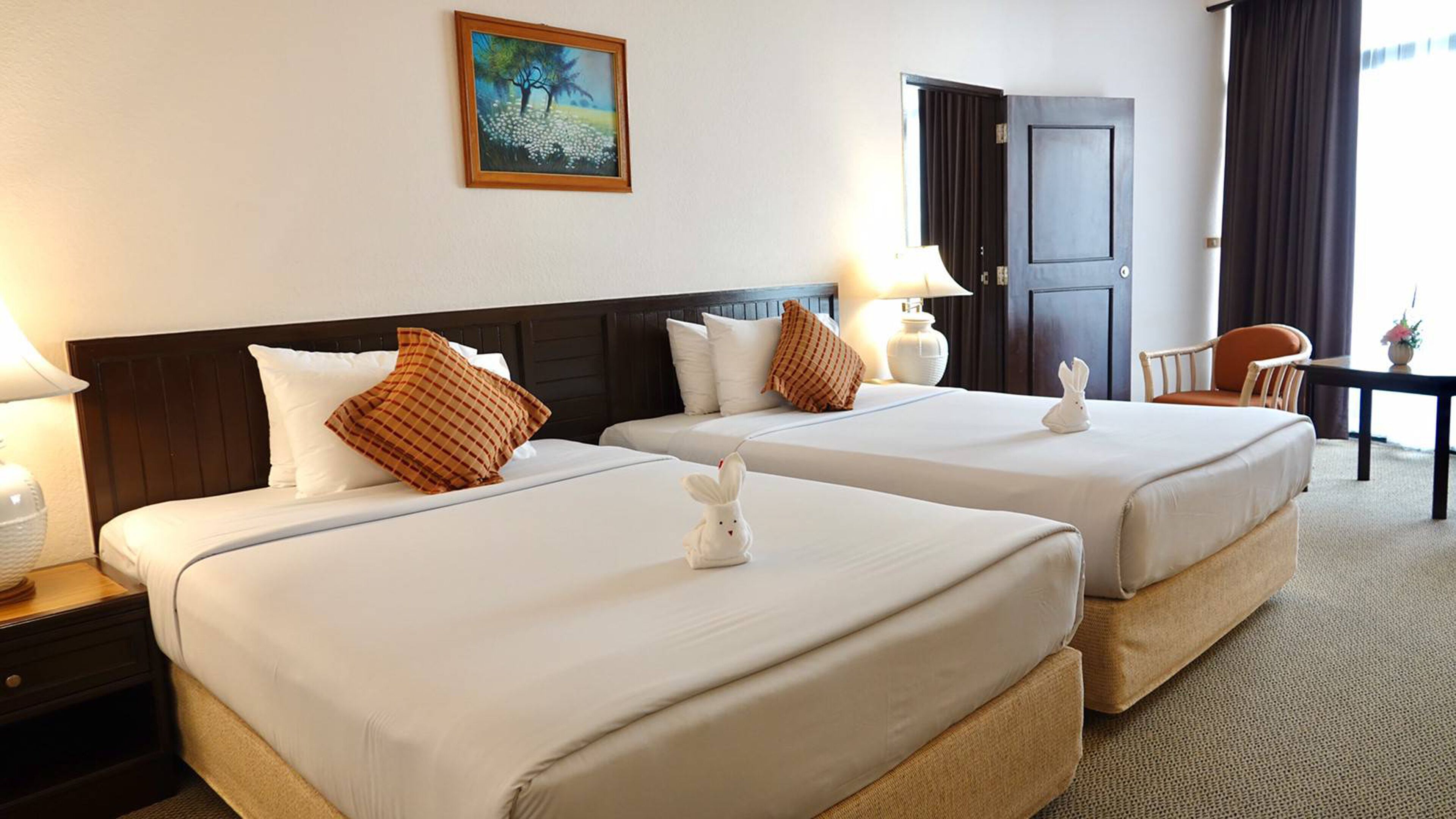A hotel room with two beds, a painting on the wall, a desk with a chair, and a door.