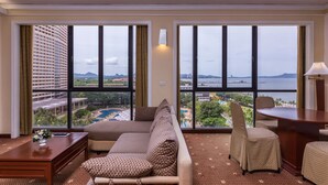 A hotel room with a large window offering a view of a cityscape and a body of water. There is a sofa, a chair, and a small table with a lamp. - Ambassador City Jomtien Pattaya - Ocean Wing (Sattahip)