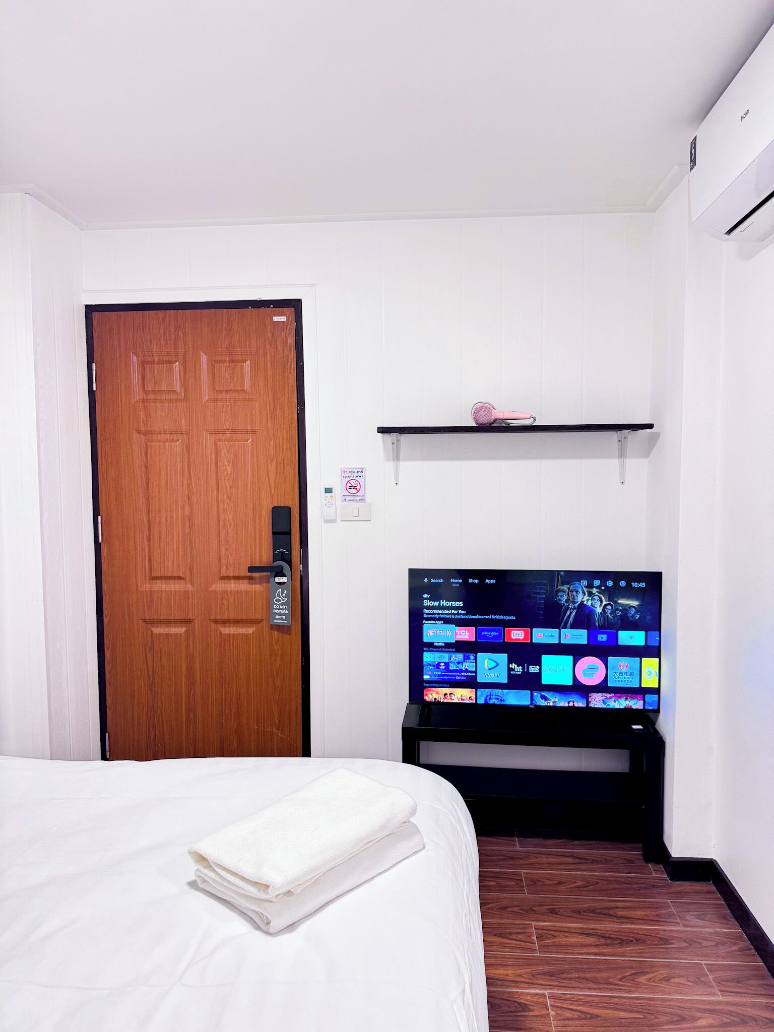 A hotel room with a bed, a television displaying various channels, a wooden door, and a shelf with a pink object.