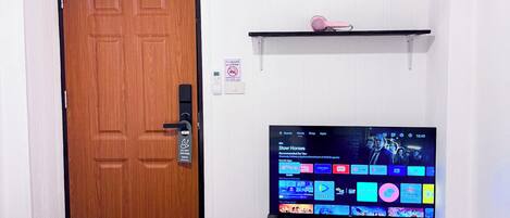 A hotel room with a bed, a television displaying various channels, a wooden door, and a shelf with a pink object.