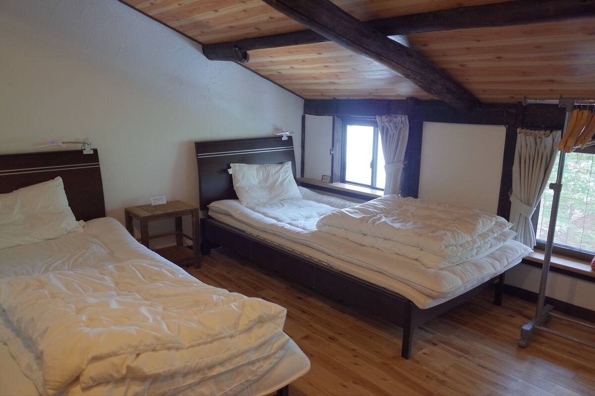Una habitación con dos camas, suelo de madera, una ventana con cortinas y techo de madera.