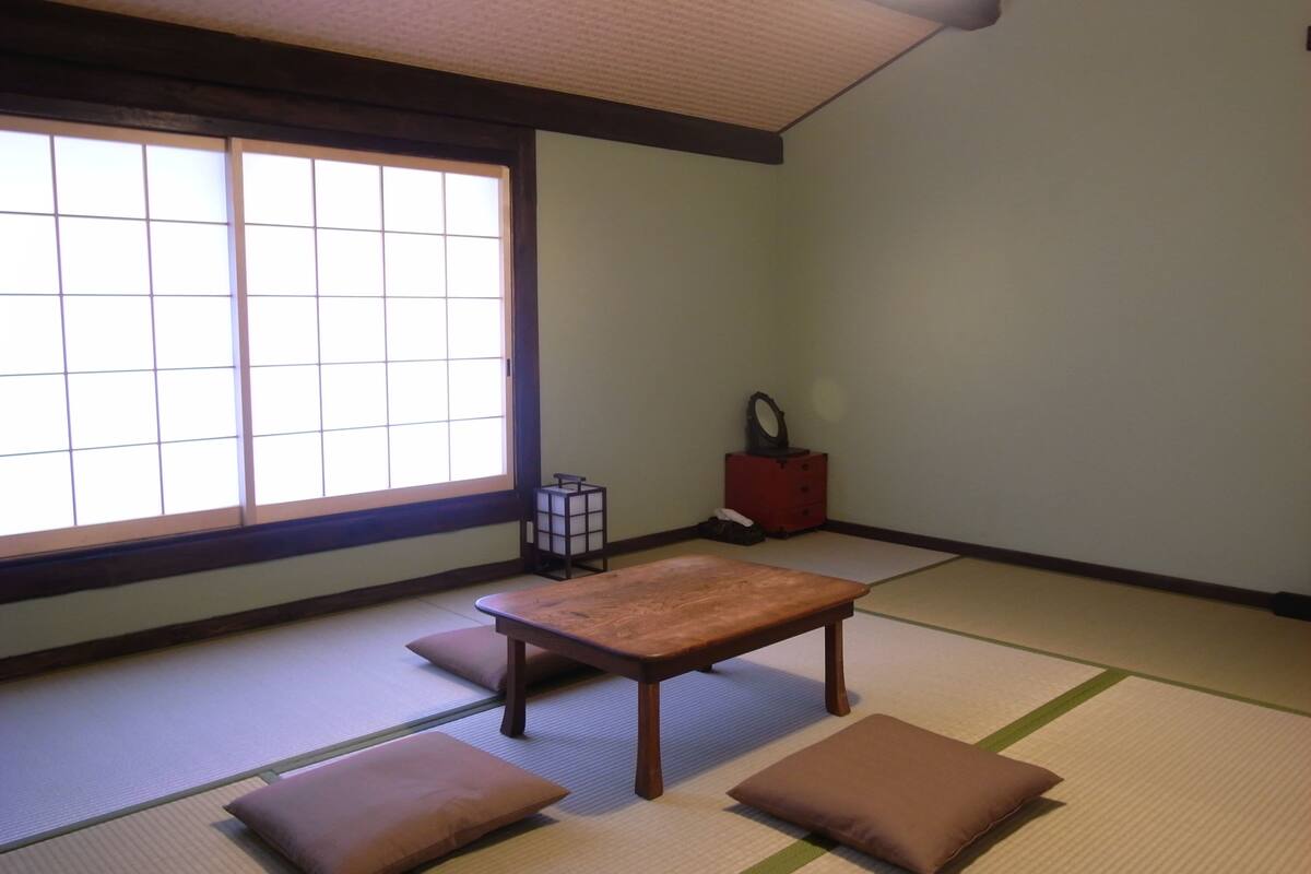 Una habitación con esteras de tatami, una mesa de madera baja y cojines, un armario pequeño y una ventana con vistas.