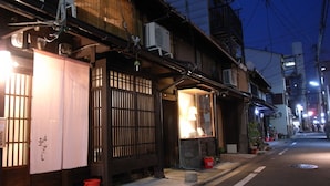 A traditional Japanese building with a wooden gate, a small shop with a lit window, and a street with buildings and overhead wires. - Guesthouse Chikayoshi (Kyoto)