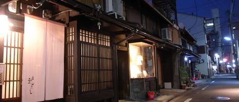 A traditional Japanese building with a wooden gate, a small shop with a lit window, and a street with buildings and overhead wires.