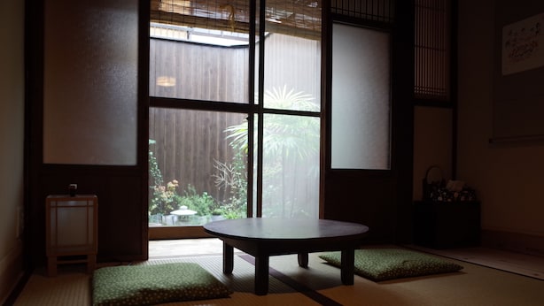A tatami mat room with a low table and cushions, large windows with a view of greenery outside. - Guesthouse Chikayoshi (Kyoto)