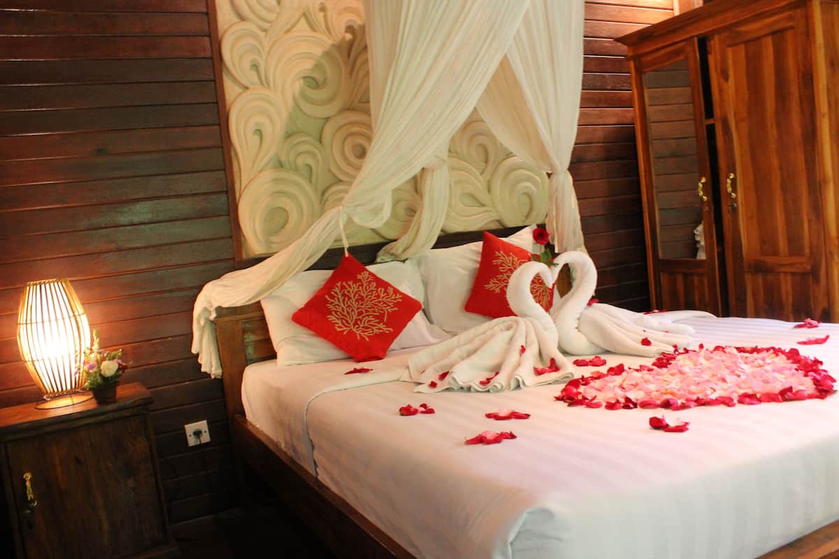 A neatly made bed with a canopy, red decorative pillows, and rose petals scattered on the bed.
