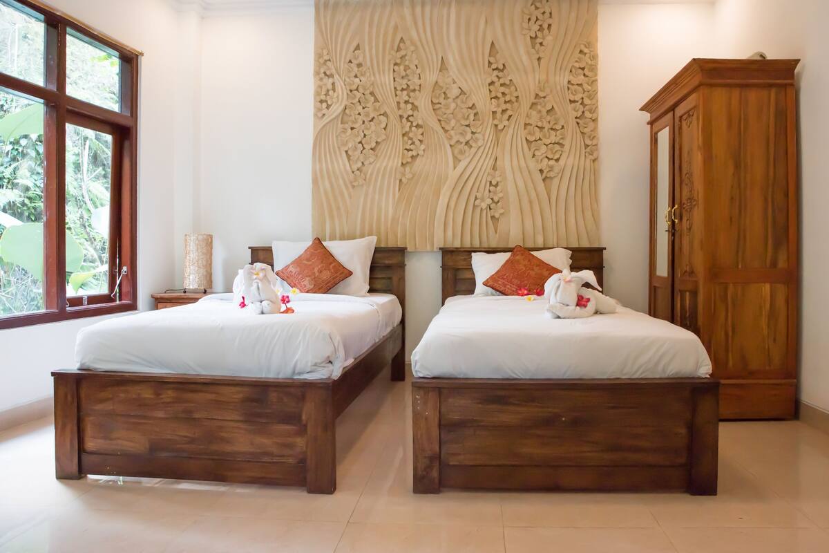 A room with two wooden beds, a decorative wall, a wooden wardrobe, and a window with a view of greenery.