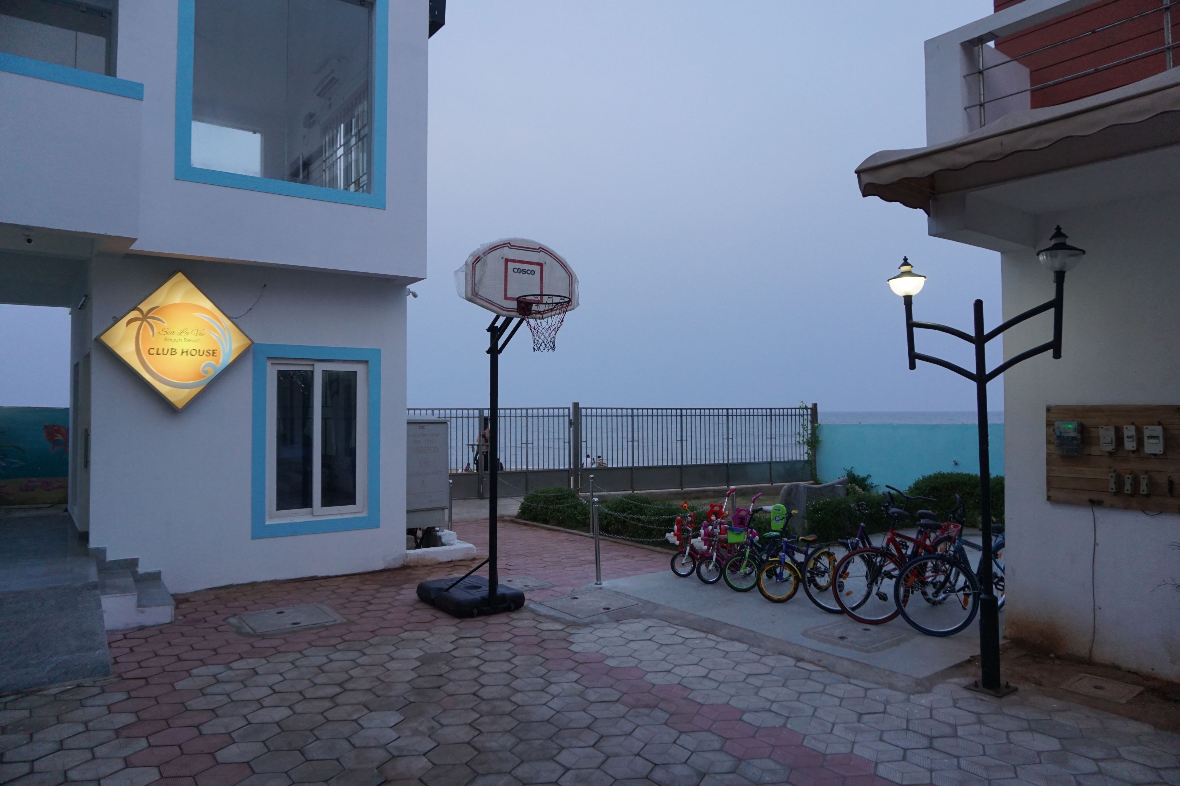 Een basketbalveld met een basketje, een fietsenrek en een gebouw met een bord waarop staat 'Sun Club House'.