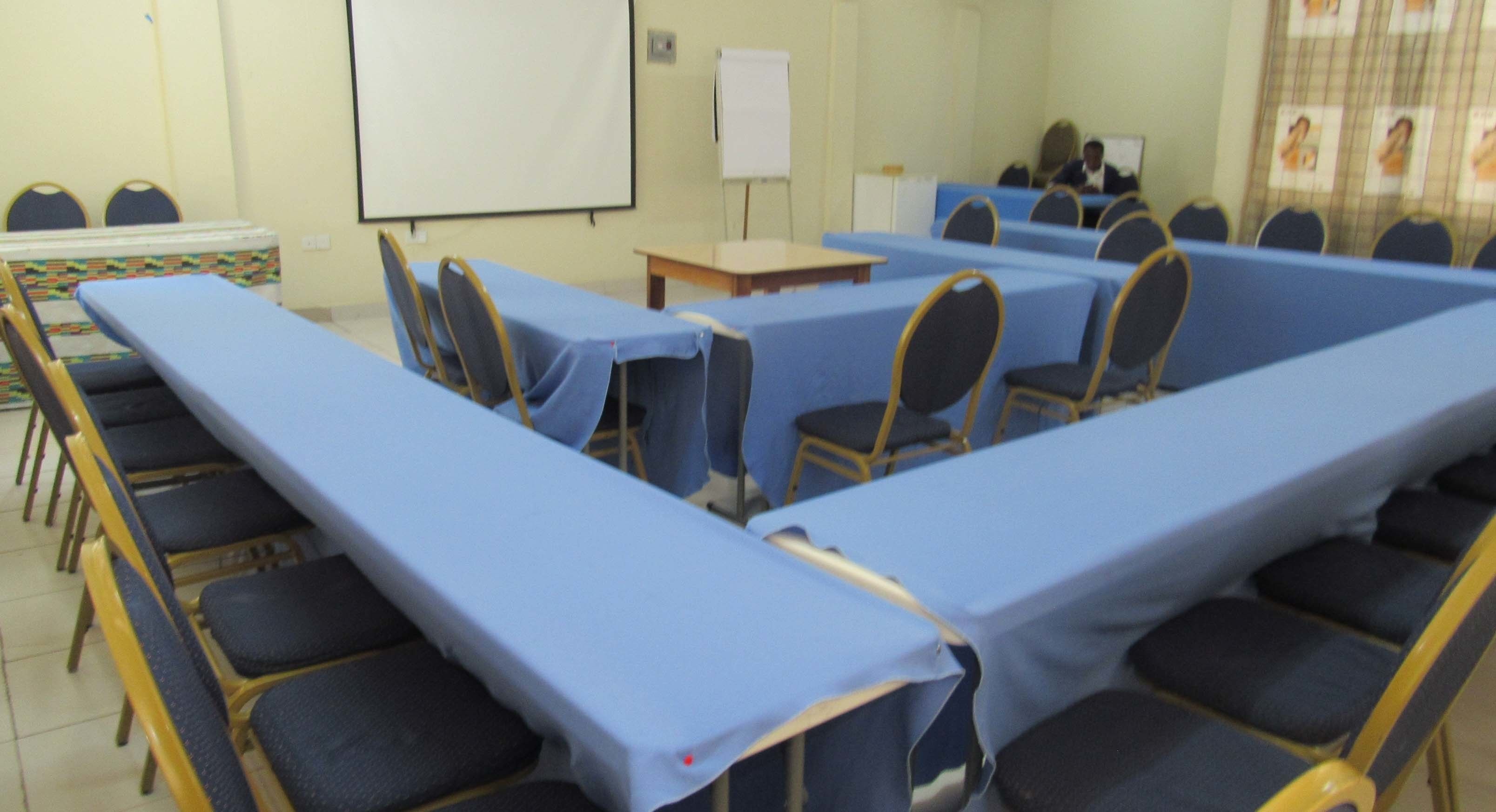 A conference room with long tables and chairs, a whiteboard, and a projector screen.