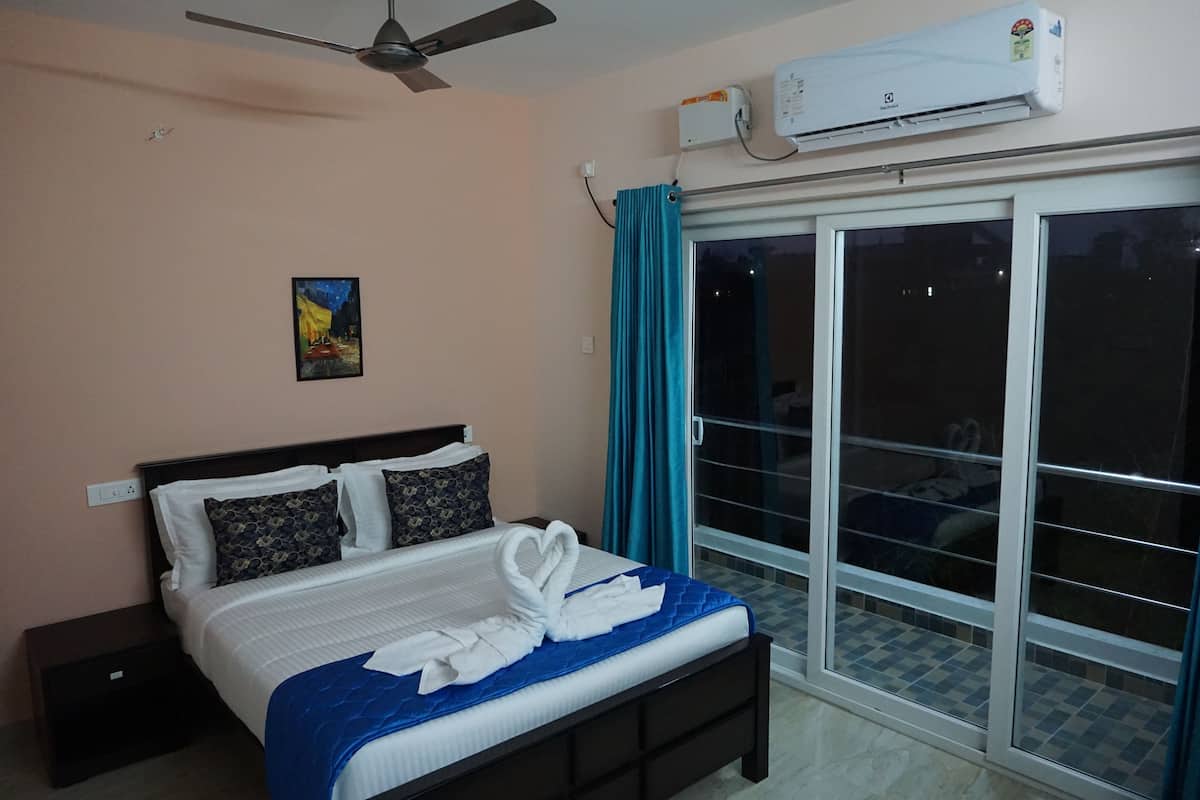 A hotel room with a bed, a ceiling fan, an air conditioner, a painting on the wall, and a view through a glass door.