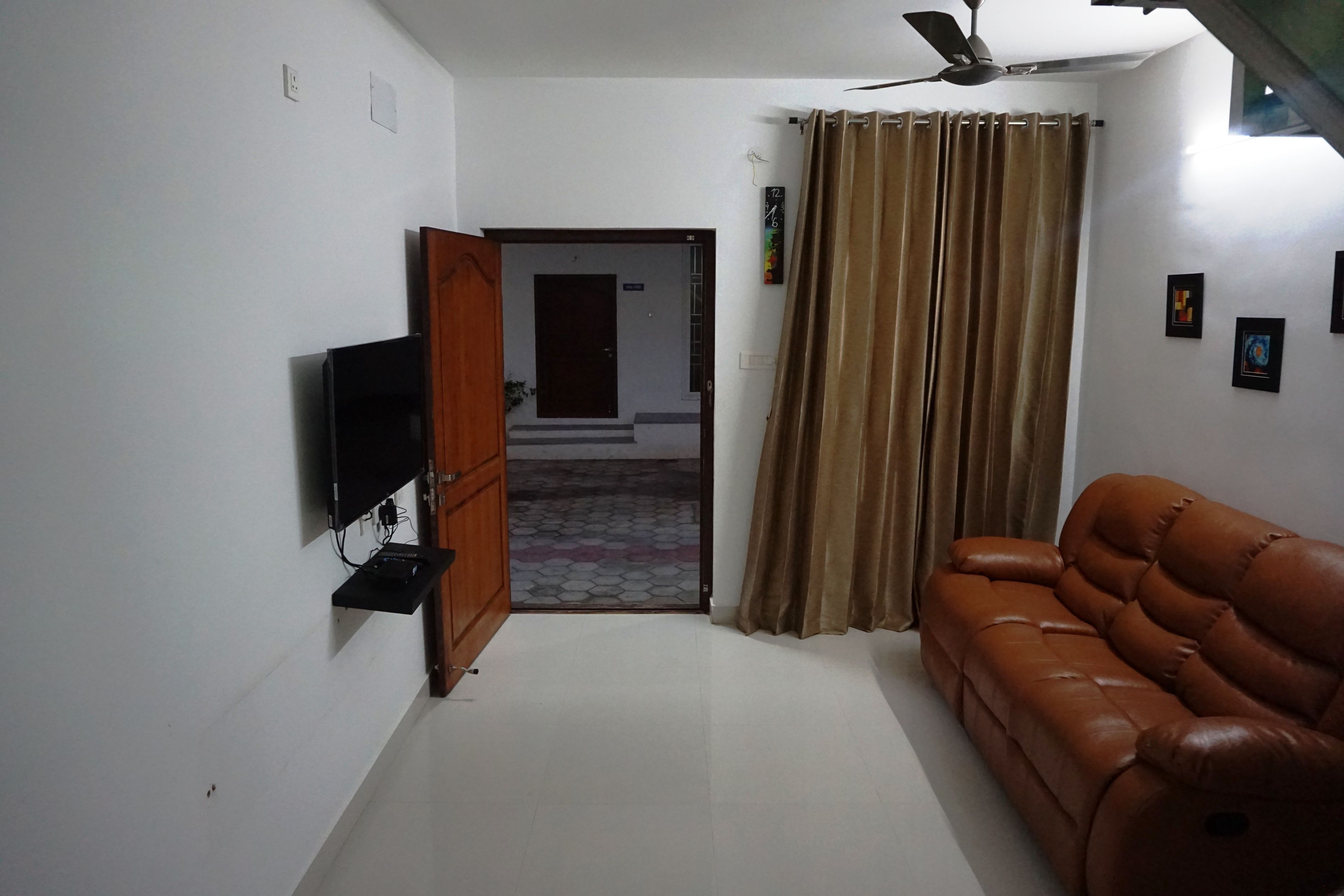 A living room with a brown leather sofa, a television mounted on the wall, and a ceiling fan.
